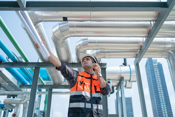 A construction engineer in a safety vest and helmet inspects industrial pipes on a rooftop while...