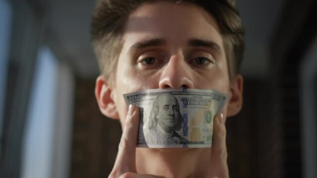 A young man covering his mouth with a hundred dollar bill, closeup. Concept: blackmail, bribery, censorship, bribe, corruption