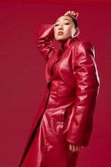 Fashion asian brunette in red leather coat posing against red background with hands on hips and head tilted to the side