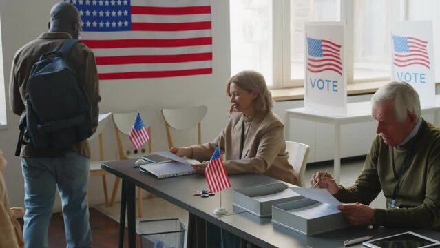 Zoom off shot of senior election inspectors sitting at table, welcoming multi-ethnic voters and giving them ballots at polling place