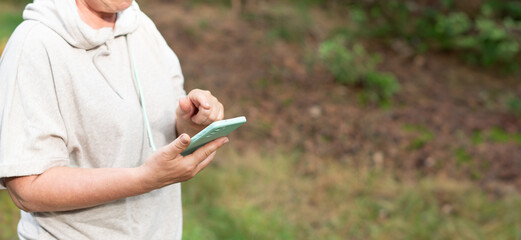 woman in the forest in beige knitted suit performs her daily requirement of steps, inhaling the air in a pine forest, looks at the route map on her smartphone and starts the pedometer