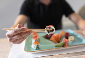 Close-up shot of a persons hand using chopsticks to hold a single sushi roll over a plate of...