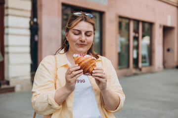Beautiful overweight brunette 30s girl posing on the street, she has big croissant meal in hands....
