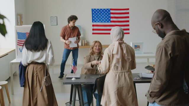 Diverse voters queuing in line, talking paper ballots from election inspector and walking to voting booths at polling place