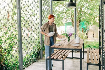 Beautiful adult redhead woman setting table for dinner on the fresh air on the terrace dressed casual fit eco style