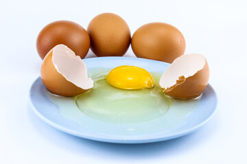 a cracked raw egg on a small bluish plate and three eggs next to it on a white isolated background