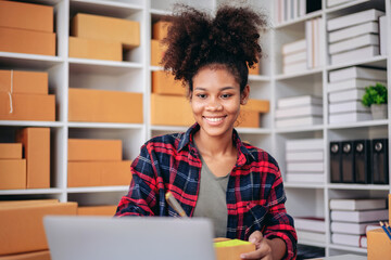 A young woman is sitting at a desk with a laptop and a stack of boxes behind her