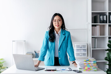 A woman in a blue suit stands in front of a desk with a laptop