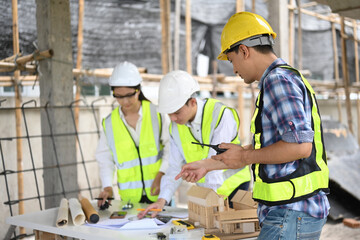 An architect, engineer, and foreman discuss plans at a construction site. They review blueprints and models, wearing safety gear. The background shows scaffolding and building materials