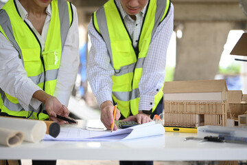 Two professionals examine blueprints and a scale model at a construction site. They point out details, surrounded by construction materials and equipment