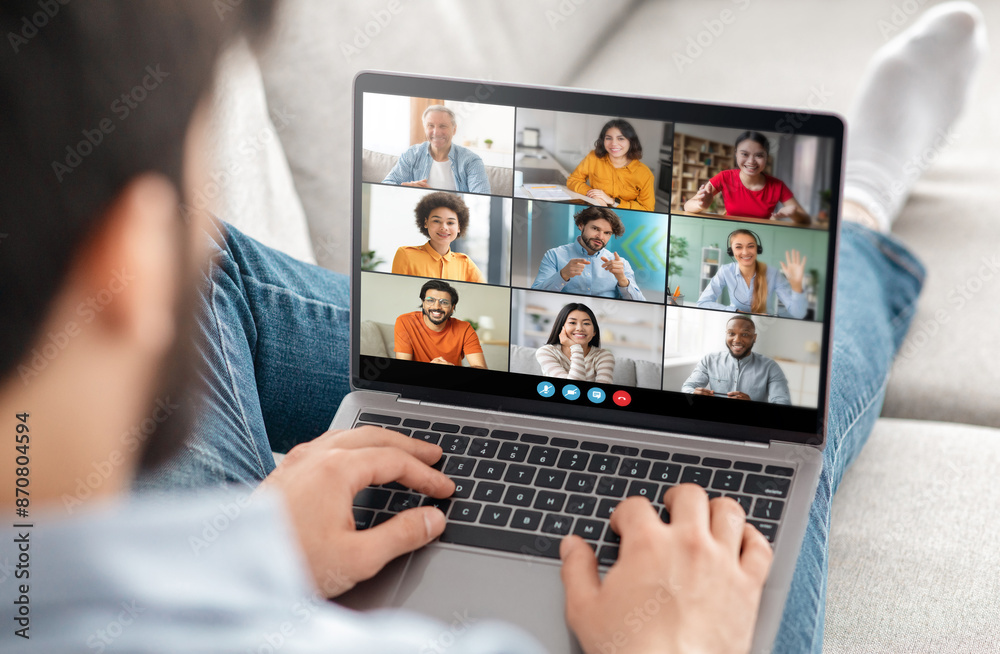 Poster A man is sitting on a couch at home, using a laptop to participate in a video conference call with several other people. He is typing on the keyboard and appears to be actively engaged