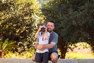 A Brazilian photographer father teaching his young son how to use a camera in a lush, green area. The father helps him frame the shot, capturing a bonding and educational moment on Father’s Day