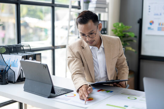 A man in a tan suit is sitting at a desk with a laptop and a clipboard
