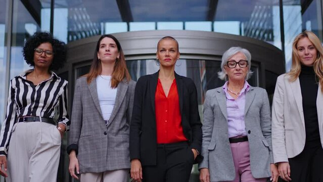 Low angle team of successful and empowered formal business women walking towards camera in front of entrance of modern office building . Confident adult diverse professional females partners outdoors