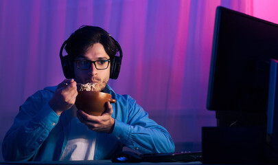A young man sits in front of a computer screen, wearing headphones, while eating a bowl of noodles....