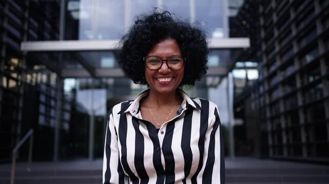 Camera zoom of adult African American business woman posing smiling standing with hands clasped together in front office building. Cheerful and empowered black female looking happy at camera outdoors
