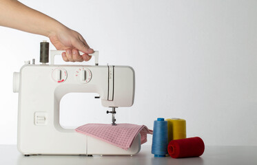 The hand of a seamstress holds a modern automatic sewing machine on a white background with multi-colored spools of thread. Copy space for text. Tailoring services