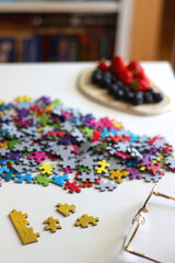 Colorful jigsaw puzzle pieces, cup of tea or coffee, eyeglasses and fresh berries for snacking. Hygge at home. Selective focus.