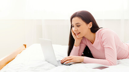 A woman is laying comfortably on a bed, focused on using her laptop computer. She appears relaxed and engaged in her online activities, websurfing, copy space