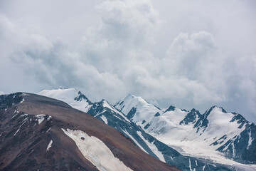 Dramatic view to large rock mountain range with snow-capped pinnacle in gray cloudy sky. Atmospheric landscape with snow cornice on snow-white pointy peak. Big rocky mountains and snowy peaked top.