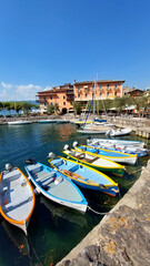 Obraz premium Colorful boats docking at a pier on a sunny day with a picturesque village in the background at lake, Garda