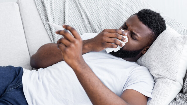 African American man lies on a couch, holding a thermometer in one hand and a tissue to his nose in the other. He appears to be sick.