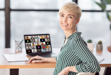 Mature woman with short blonde hair sits at a desk in an office setting with a laptop open in front...