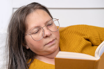 mature woman 50 years old in yellow sweater and glasses semi-reclined, reading Interesting book, joy and importance of literature in one's life