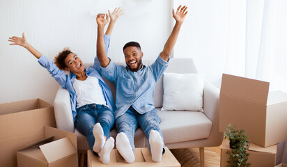 Housewarming Party. Happy Afro Couple Raising Hands Smiling At Camera Sitting On Sofa Among Moving Boxes In Own Flat.