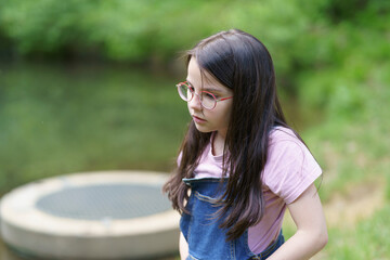 A little cute teenage girl in glasses stands near a pond. Child in denim overalls. Happy childhood...