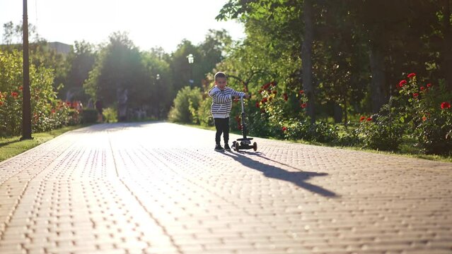 Cute smiling baby boy turns his scooter on the paved road in the park. Kid walks approaching camera and then rides pushing from the ground.