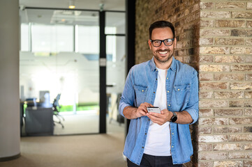 Portrait of happy young man developing mobile phone applications while leaning on brick wall in office