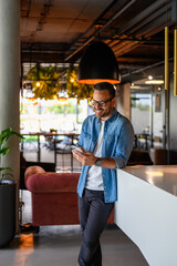 Smiling male entrepreneur reading online messages over smart phone while standing in modern office