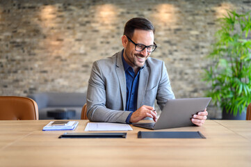 Handsome IT engineer in eyeglasses smiling and developing website over laptop on desk in modern...