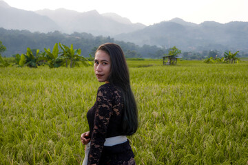 a beautiful Indonesian woman wearing a black Balinese kebaya with a background of rice fields and mountains