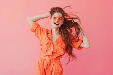 A joyful woman in orange jumpsuit and sunglasses, laughing with wind-blown hair against a pink background.