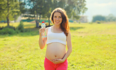 Obraz premium Pregnancy, happy young pregnant woman drinking water from plastic bottle in sunny summer park