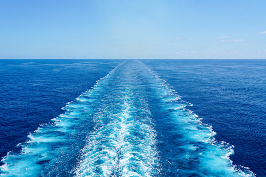 Wide turbulent white foam water trail behind a large cruise ship in a calm blue sea on a sunny day