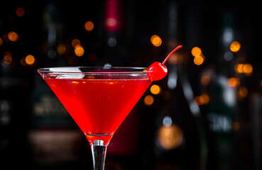 Red alcoholic cocktail drink with cherry on dark background, bar counter with bottles, close-up