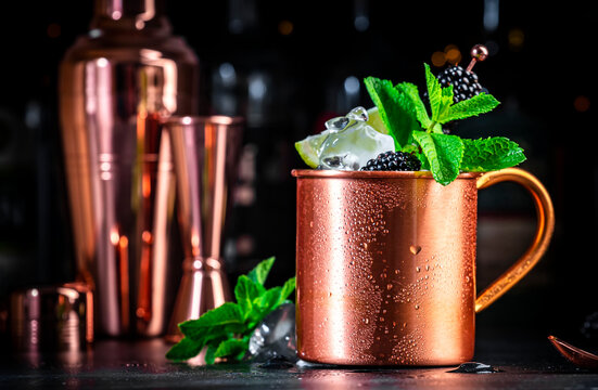Blackberry Moscow mule alcoholic cocktail drink in copper mug with lime, ice, ginger beer, vodka and mint. Black bar counter background, bar tools, bottles