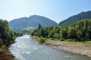 Clear flowing of Cheremosh River flanked by lush green trees winds through stone banks, and in the background, Carpathian Mountains under a bright blue sky