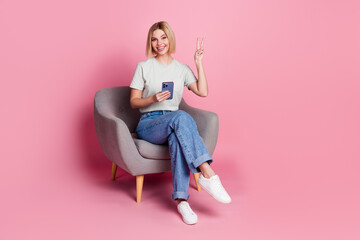 Photo of cheerful lovely nice girl wear stylish grey clothes sit chair hold device v-sign isolated on pink color background