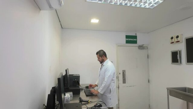 A dark-skinned Latino male with a large nose and a thick beard who wears a white coat and interacts with computer equipment while carrying out scientific activities.