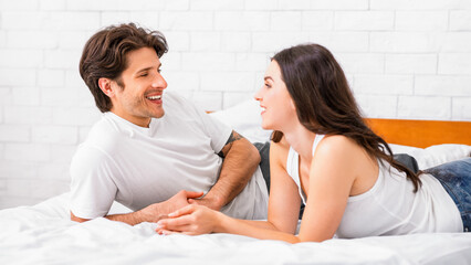 A young couple lies on a bed together, facing each other, engaging in a lighthearted and intimate conversation. They are both smiling, suggesting a sense of joy and affection between them