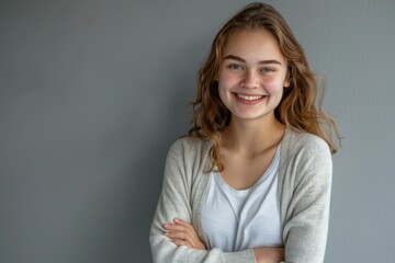 Obraz premium A closeup portrait of a smiling young woman with her arms crossed in a confident stance, standing against a neutral grey backdrop