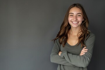 Obraz premium A portrait of a smiling young woman with long brown hair, wearing a grey sweater, posing with her arms crossed against a plain grey background