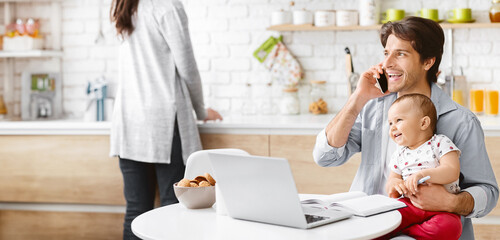 A man sits at a kitchen table with a laptop in front of him. He is holding his baby in his lap while talking on his phone. The baby is smiling and looking at the camera