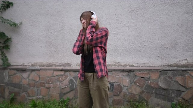 young woman in red plaid shirt and beige pants dances hip-hop while listening to music through large white headphones. She stands against gray wall with stone base, smiling and enjoying the rhythm