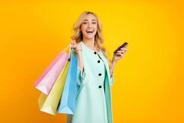 A young woman with long blonde hair, wearing a light blue coat, smiles excitedly while holding a smartphone and several brightly colored shopping bags in front of a vibrant yellow background.