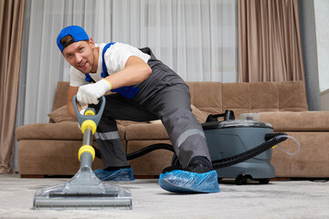 An efficient cleaner from a top cleaning firm, donned in special attire with cap and shoe covers, effectively vacuuming a carpet in a generously sized living area.